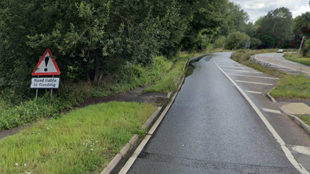 A triangular road warning sign on a verge beside the A33 dual carriageway at the crash scene says "Road liable to flooding"