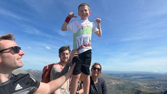 Oscar is standing at the summit of Snowdon. A man wearing sunglasses is holding his leg, whole a man and a woman stand behind him, also holding onto him.
