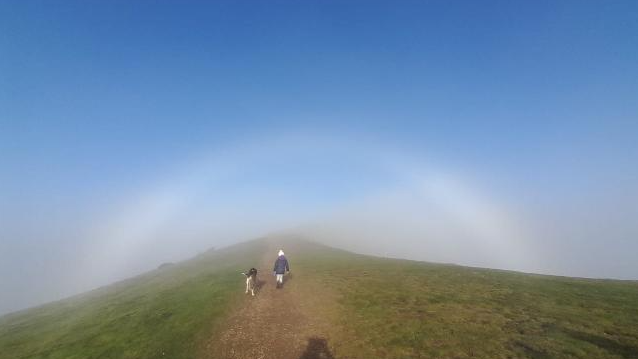 A person's shadow shows them taking a photo of another person walking a dog on the hills with blue sky around them.