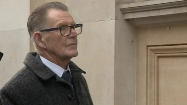 Patrick Devaney outside Bristol Crown Court. He is side-on to the camera and wears a dark grey coat, white shirt and dark blue tie. He has grey heair swept back and large dark-framed glasses. 