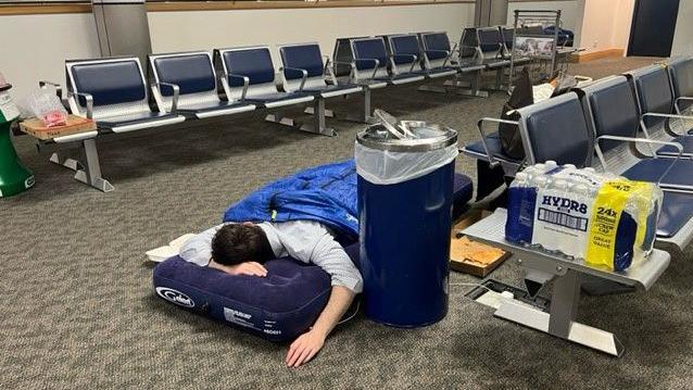 A person is lying in a sleeping bag on an inflatable bed in an airport waiting area with a pack of bottled water and a rubbish bin next to them.