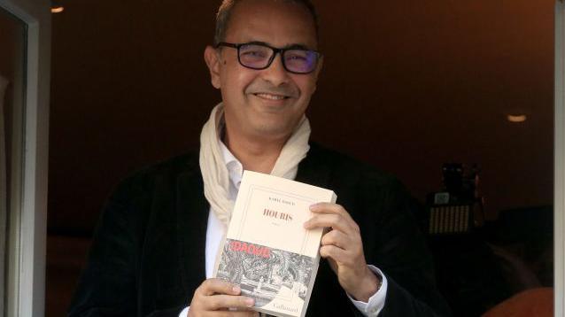 A man, Kamel Daoud, in black-framed glasses smiles as he stands in an open window, holding a white copy of his prize-winning book. He is wearing a dark blazer and a white shirt, with a cream-coloured scarf lightly looped around his neck.