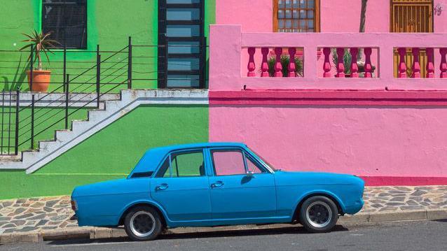 A bright blue car stands in front of neon green and pink buildings.
