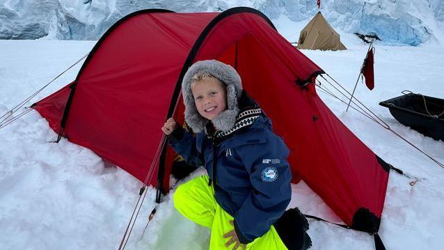 Jamie with blond hair and smiling wearing a furry grey hat, blue coat and lime green trousers sitting in snow kneeling down in front of a tent in Antarctica 