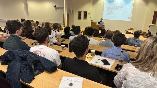 People on tiered seating looking down at a speaker with a projection behind him