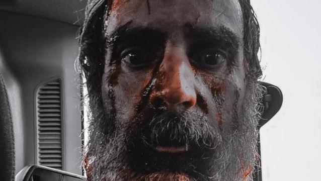 A close-up of a man with staring eyes and facial hair. His head is smeared in blood.