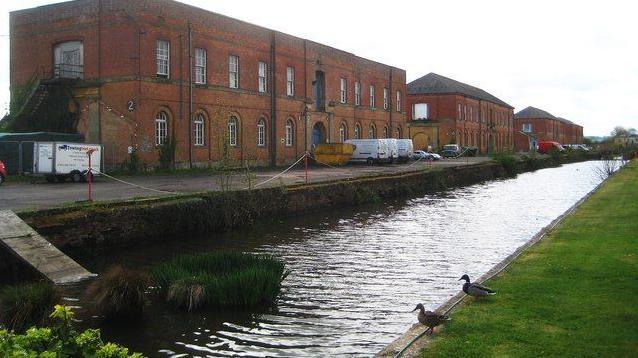 A section of a canal with two ducks watching the water from the bank in the foreground. There are two-storey brick barrack buildings to the left.