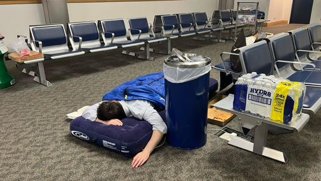 A man is laying on an air mattress in the terminal waiting area, surrounded by seating. 