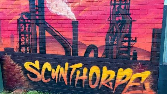 A mural of the steel works in black on a wall with a red background and the word Scunthorpe in yellow and orange graffiti style text.