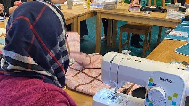 A view of Aziza from behind sitting on a chair behind a desk with pink material in front of a sewing machine.