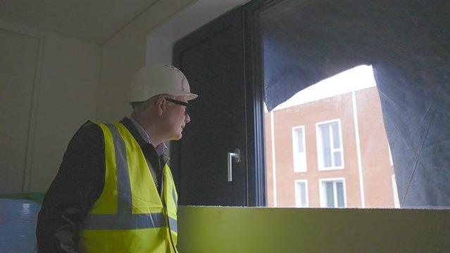 Mayor Richard Parker stands inside one of the new social homes under construction at Port Loop. He is looking out of a window at other buildings in the development. The room he is in is still being built. He wears a white hard hat and black rimmed glasses with a green high-vis vest over a dark suit jacket.