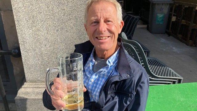 Bjorn has short grey hair and is clean shaven. He wears a light blue and white checked shirt and a blue zip-up jacket. He is holding glass of beer.