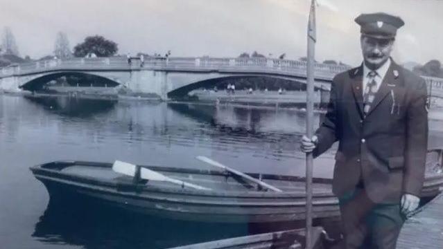 A black and white photo showing a man wearing a dark uniform and peaked cap. He is holding a large boating pole and standing in front of rowing boats at a large lake in a park. In the background a double-arched, stone footbridge crosses the lake.
