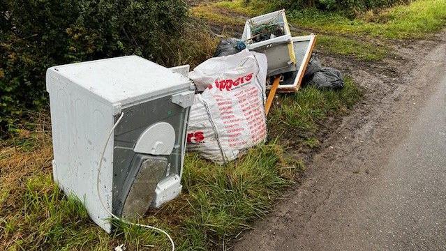 The verge of a country lane near a gateway with a fridge, a builders bag and other rubbish lined up