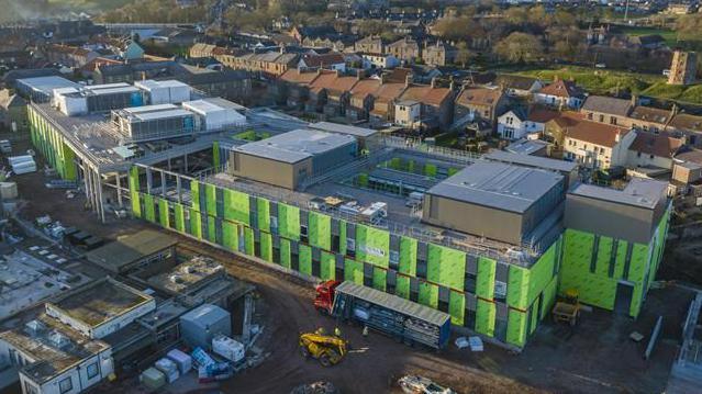 An aerial view of the new Berwick hospital mid construction. Green panelling has been paced over the sprawling hospital building while work is under way. An excavator and a large lorry are parked outside. There are rows of houses behind it.