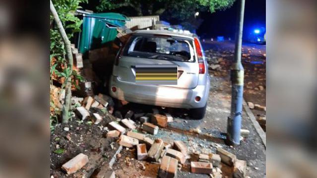 A silver car crashed with its front end into a brick wall. Surrounding the car are lots of red scattered bricks and there is also a green storage container that has been damaged