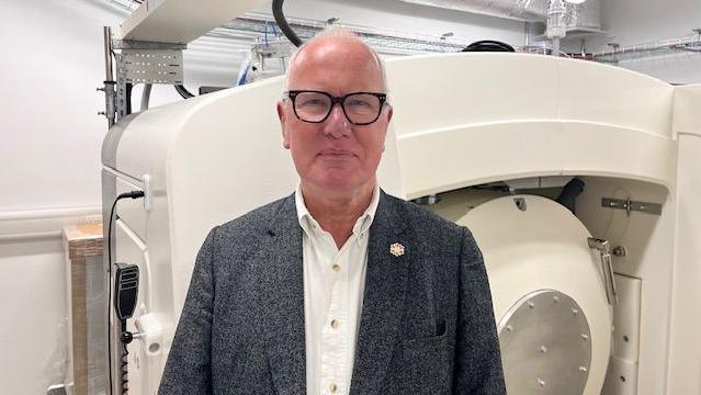 Professor Nick Stafford stands in front of new medical equipment at Castle Hill Hospital. He is wearing a grey blazer and a white shirt. He has black glasses and a bald head