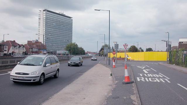 Photo taken in 2005 of a main road with cones and cars on it. There is a yellow fence to the right of the image. 