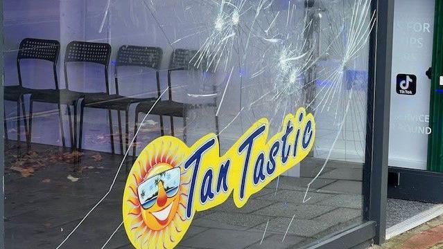 Badly cracked shopfront window with Tan Tastic blue and yellow logo with sun wearing shades 