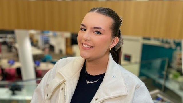 Mollieanne wearing a white jacket and black top standing in a library with books and people sitting at desks in the background