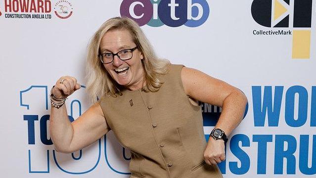 Jane stands in front of a white backdrop featuring multiple sponsor logo. She is wearing a tan sleeveless buttoned vest with matching trousers and a black wristwatch. She is posing with bent arms in a flexing gesture.
