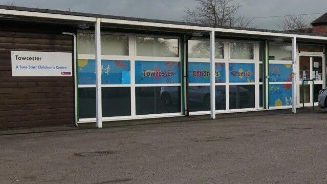 A one storey flat roof building with lots of windows at the front. A white sign that says Towcester Sure Start Children's Centre. There is a large car park in front of the building.