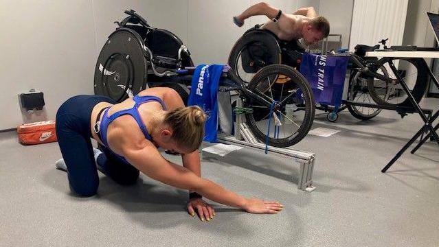 A female athlete does some stretches on the floor of a gym. Behind her, a man trains in a wheelchair.