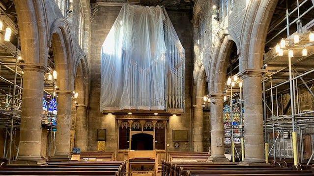 Interior of church with massive white drape covering a walled section and metal scaffolds around the sides. Pews and vaulted wooden arches in front of stained glass windows.