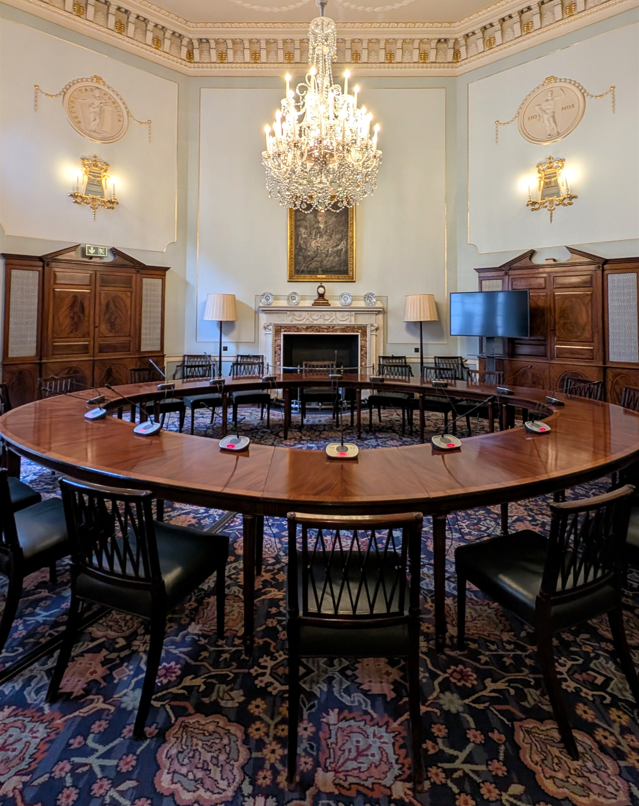 Bank of England's Committee Room with wooden chairs surrounding a round table which has microphones placed around it. The room is octagonal with blue walls, a chandelier and decorated white and gold ceiling
