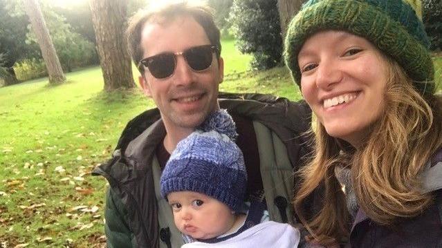 Matt and Amy Hall with baby Rory in a park. Rory is wearing a blue woolly bobble hat, while his dad has a green jacket and sunglasses. Mrs Hall is smiling into the camera and wearing a green woolly hat.