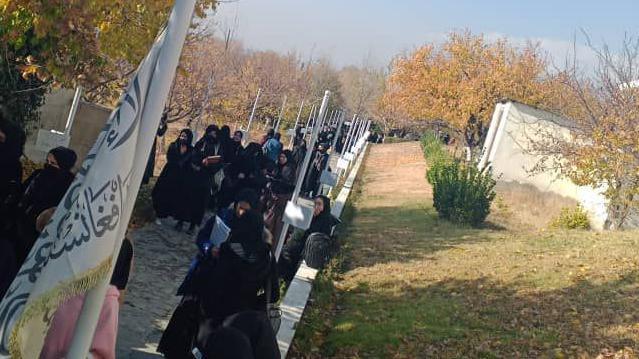 A group of women dressed in black, their hair and faces covered, walk along a path with a white flag with Arabic text on it in the foreground. The trees are autumnal.