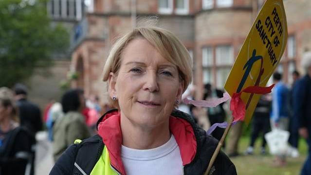 Mary Alexander has short blonde hair and holds a sign which says "Perth City of Sanctuary" outside a hotel with people in the background. She wears a black, yellow and red jacket and a white top.