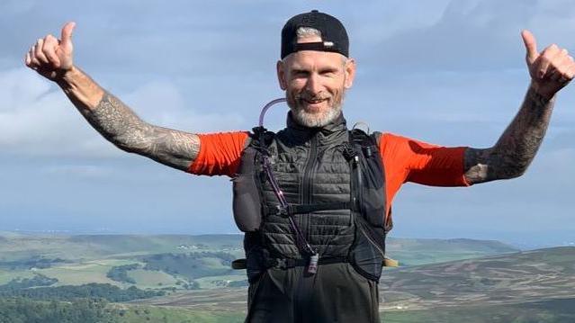 Danny Warren is pictured on a hiking trip wearing walking gear. He has both arms in the air and has his thumbs up while smiling at the camera. Rolling green hills covered in trees can be seen in the background.