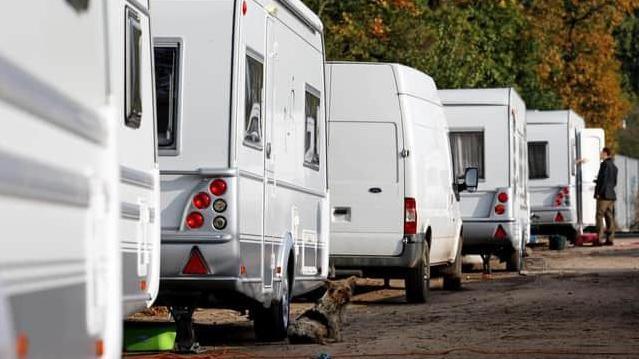 Four white caravans and a white van are parked along the edge of a muddy track. A brown dog is sat outside the door of the second caravan. A person in a black jacket and brown trousers is stood outside the door of the furthest caravan and someone's hands can be seen gesturing in conversation from inside the caravan, at the door.