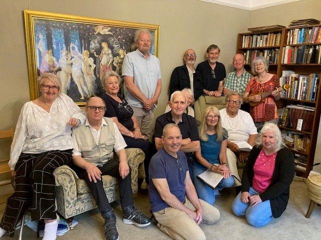Fourteen men and women, all over 50 years old, sitting in a group in a book-lined living room, smiling