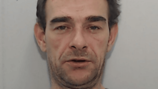 A mugshot of a middle-aged man. He has stubble, messy hair and his mouth open.