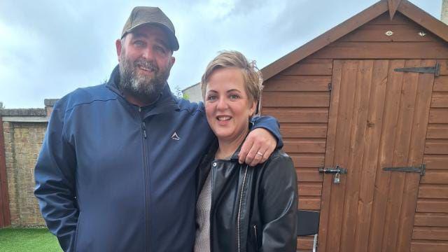 A man and a woman are standing in front of a wooden garden shed. The man is wearing a dark-blue jacket and a grey cap, and the blond-haired woman is dressed in a black leather jacket.