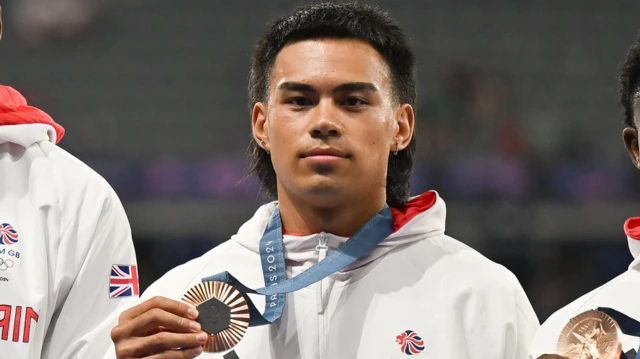 Louis Hinchliffe, wearing Team GB's white 2024 Olympics jacket, holds up his bronze medal in Paris