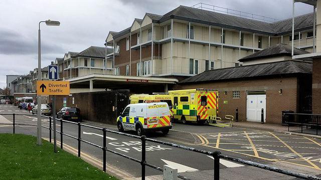 A street view of Royal Oldham Hospital. A white police van can be seen next to ambulances in the road outside.