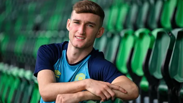 Mason Melia rests his arms on some green seats while wearing the colours of St Patrick Athletic