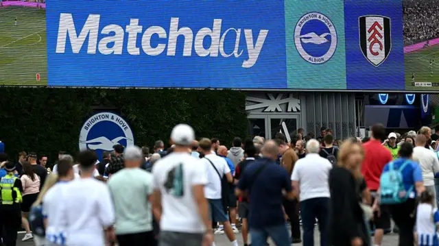 Brighton fans outside stadium where a sign shows matchday and club badges of Brighton and Fulham