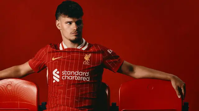 Milos Kerkez looks to the left wearing a Liverpool home shirt against a red background
