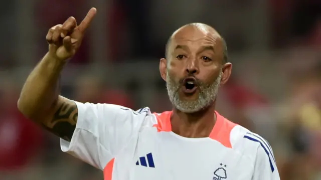 Nuno Espirito Santo holds his right arm in the air, raising his index finger, and opens his mouth while wearing a white Nottingham Forest t-shirt