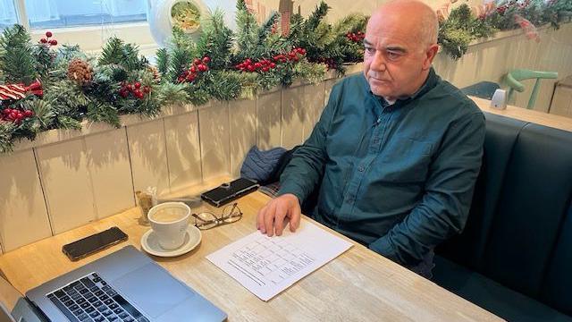 Maldwyn Pryse who has a bald head and is wearing a dark green shirt, sitting at a table with a laptop,  coffee, a mobile phone and a sheet of paper in front of him