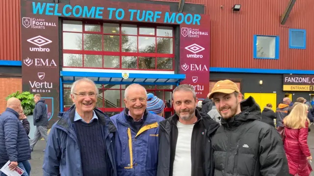 An image of four men posing for a picture in front of Turf Moor