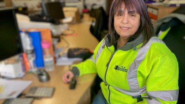 A woman in a hi-vis jacket sits at a desk 