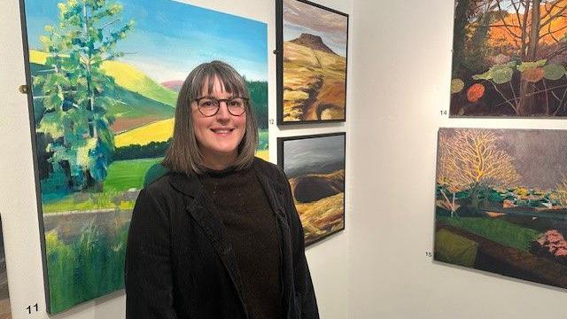 Woodhorn Museum's director of programmes and engagement, Liz Ritson. She has shoulder-length brown hair and is wearing a brown jumper and jacket and dark-rimmed glasses. She is standing in front of a collection of landscape paintings showing scenes of Northumberland.