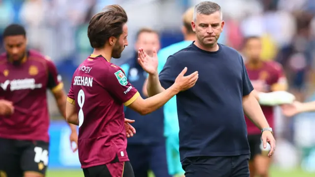 Steven Schumacher, wearing a navy blue t-shirt, claps hands with midfielder Josh Sheehan, who is wearing Bolton's maroon away kit, after the 1-0 defeat to Sheffield Wednesday in the EFL Cup at Hillsborough