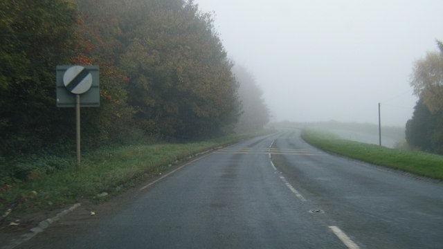 A road lined with woodland on a foggy day. In the foreground is a national speed limit road sign.