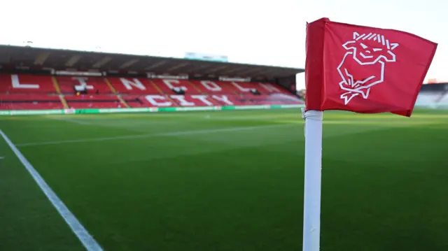 A red corner flag at Lincoln City's LNER Stadium with the club's Imp logo in white. The pitch and the red seats of the GBM Stand with Lincoln City written in white seats can be seen behind.